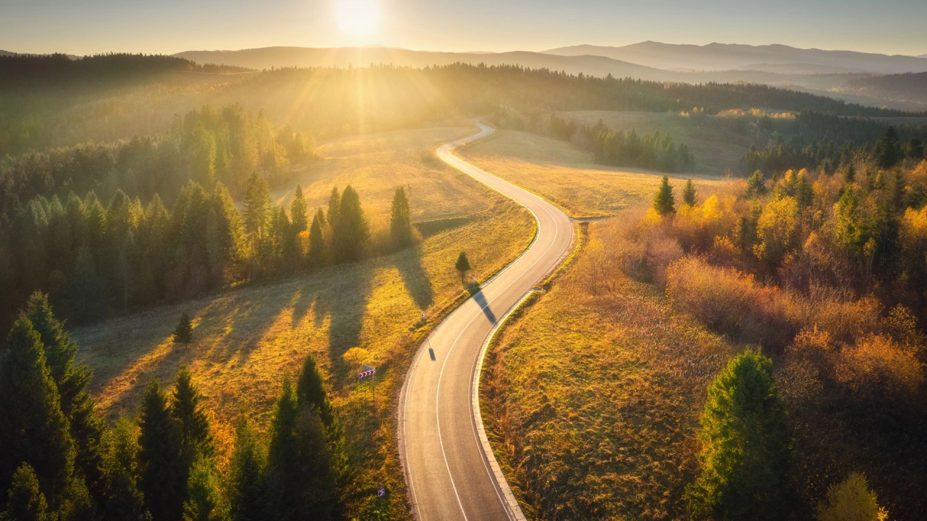 Kurvige Straße durch sonnige Landschaft mit Wäldern und Hügeln im Abendlicht.