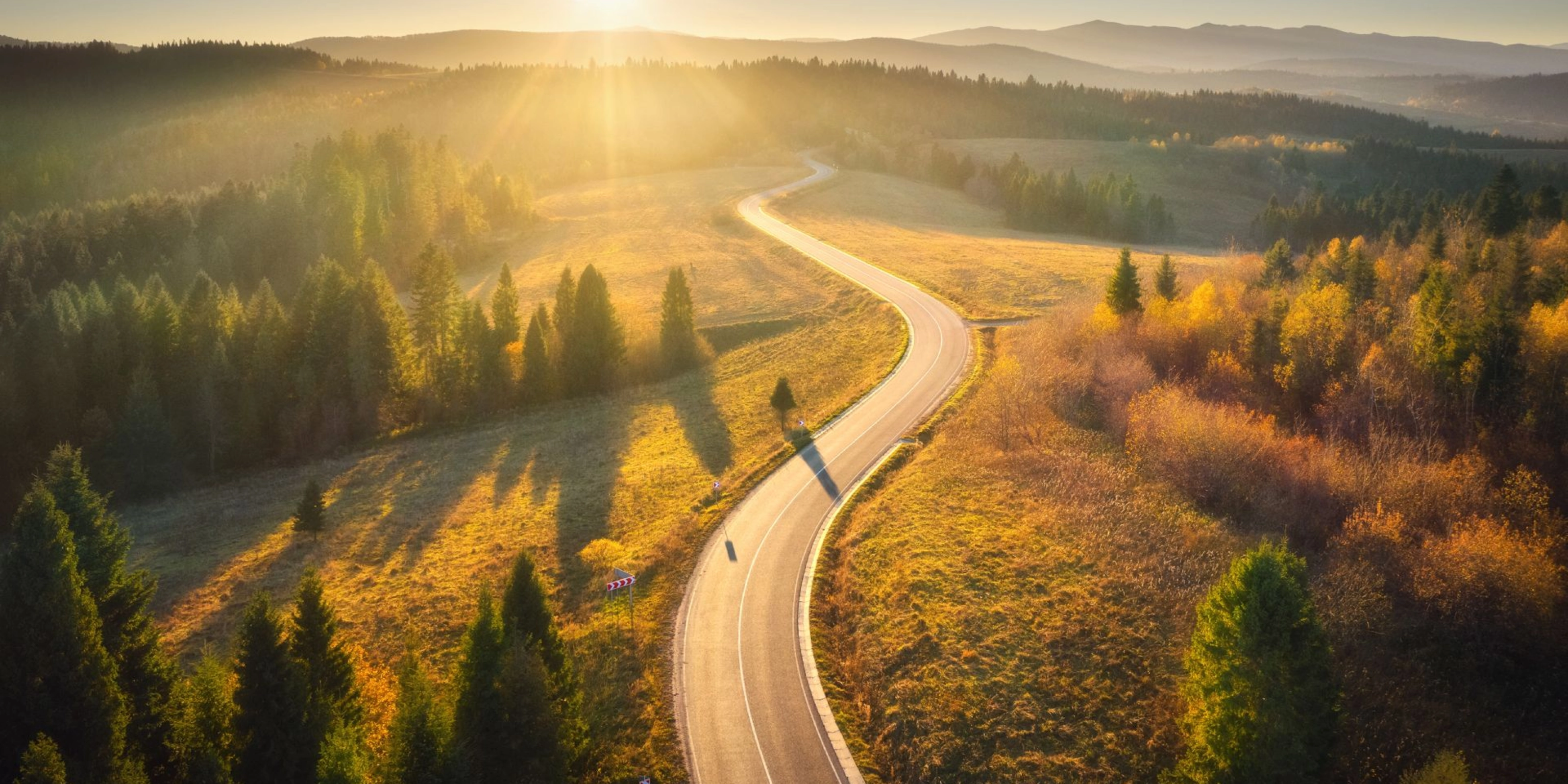 Kurvige Straße durch sonnige Landschaft mit Wäldern und Hügeln im Abendlicht.