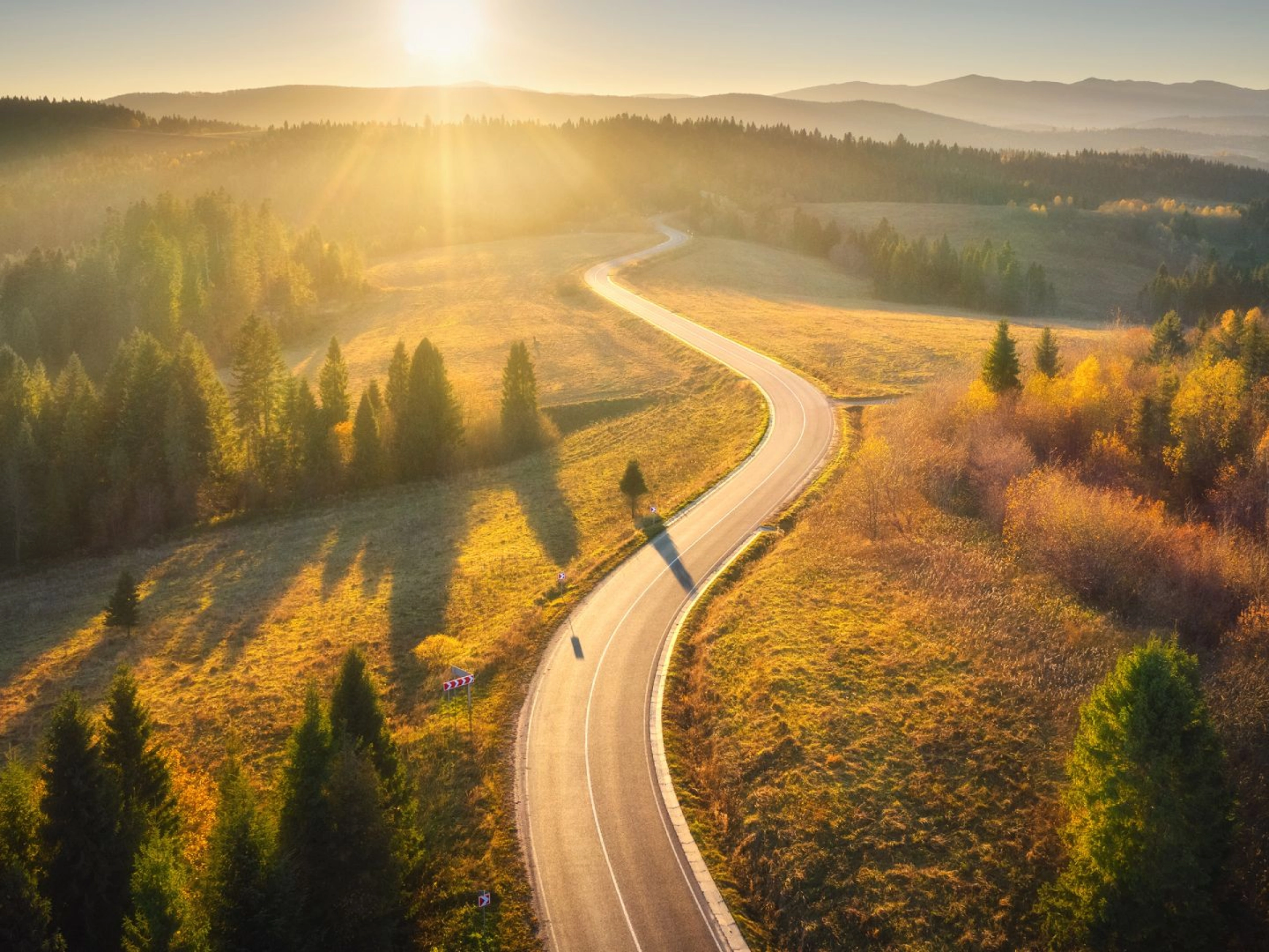 Kurvige Straße durch sonnige Landschaft mit Wäldern und Hügeln im Abendlicht.