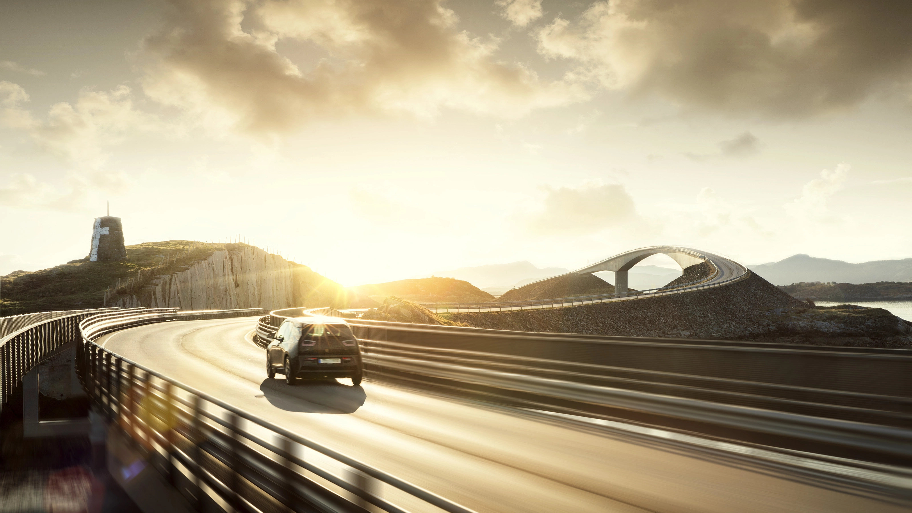 A car drives across a curving bridge at sunset in a coastal landscape.