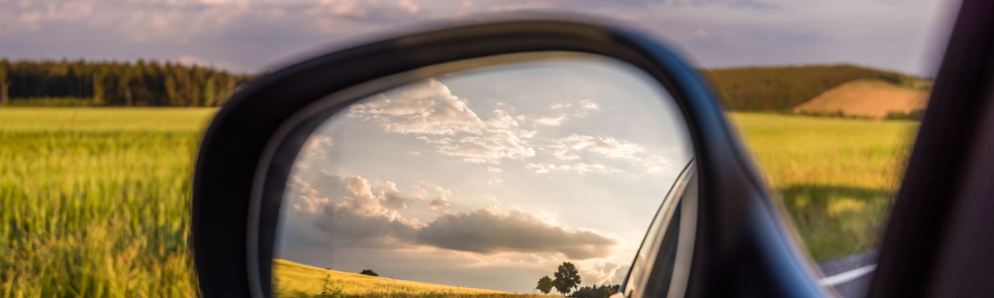 Landstraße und bewölkter Himmel spiegeln sich im Seitenspiegel eines Autos bei Tageslicht.
