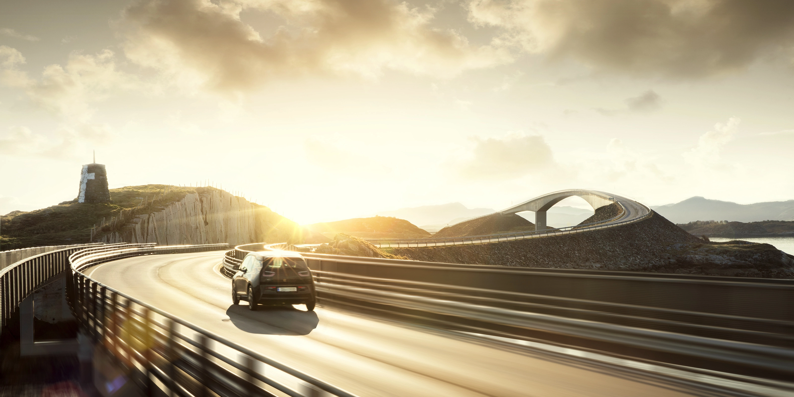 A car drives across a curving bridge at sunset in a coastal landscape.