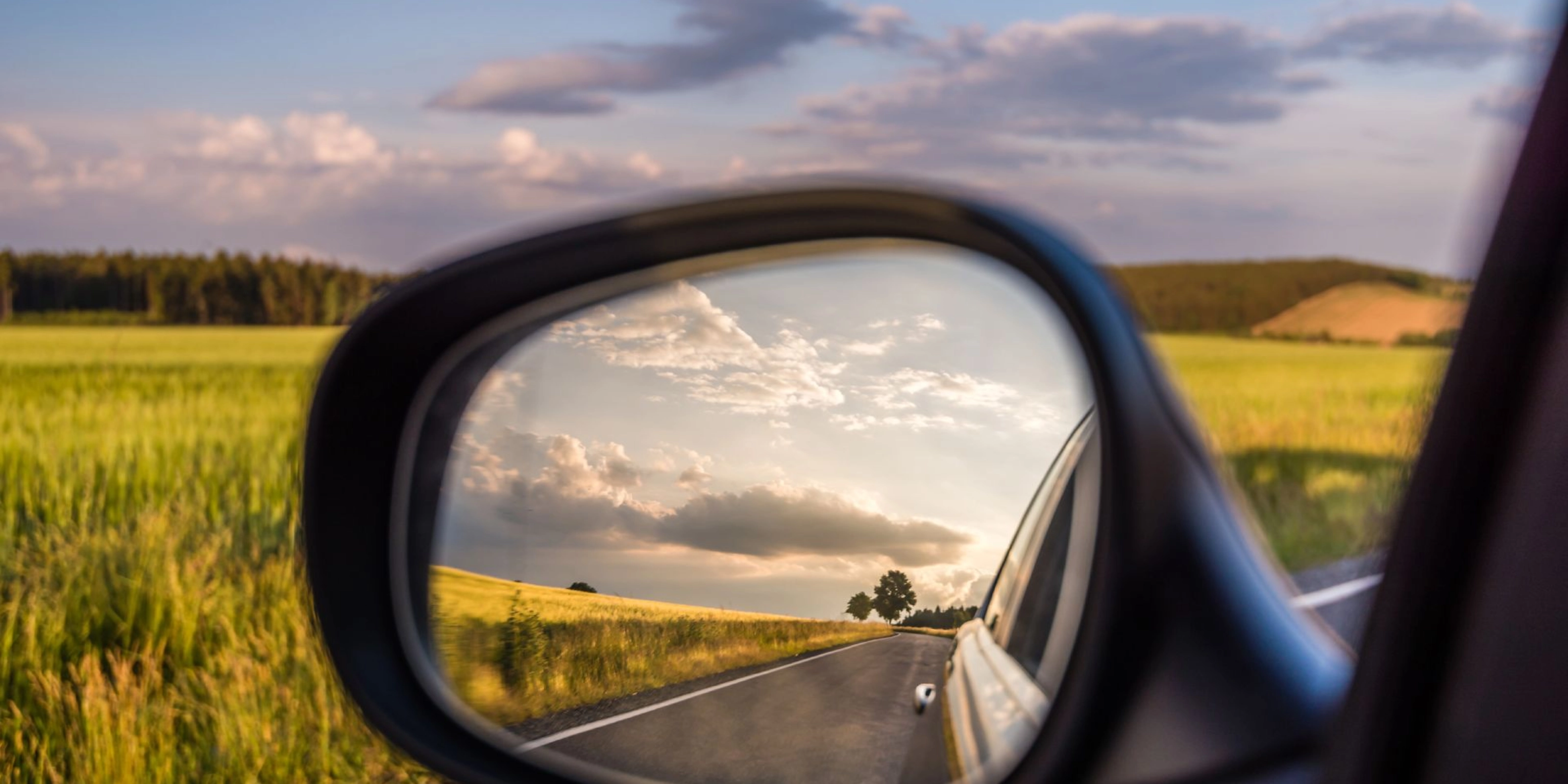 Landstraße und bewölkter Himmel spiegeln sich im Seitenspiegel eines Autos bei Tageslicht.