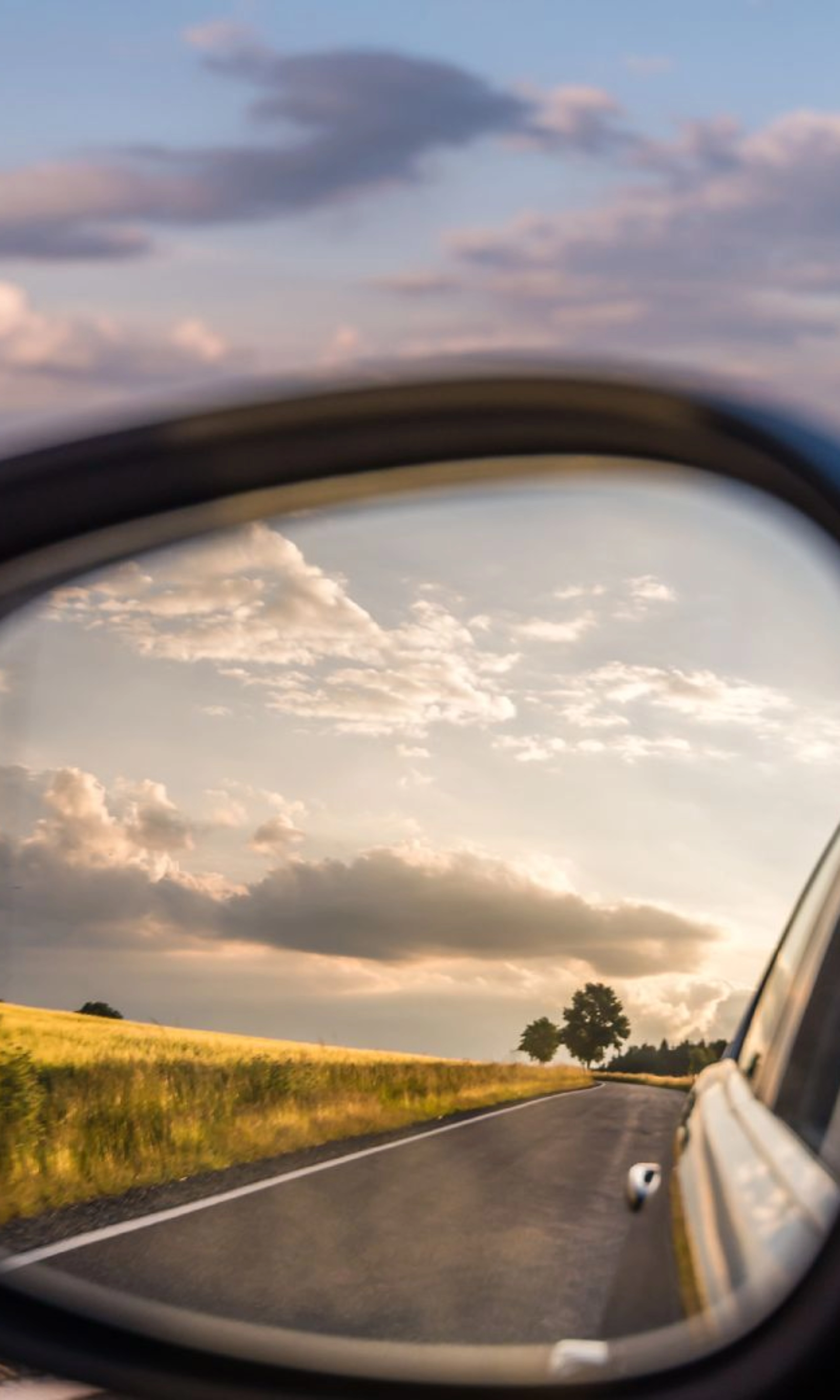 Landstraße und bewölkter Himmel spiegeln sich im Seitenspiegel eines Autos bei Tageslicht.