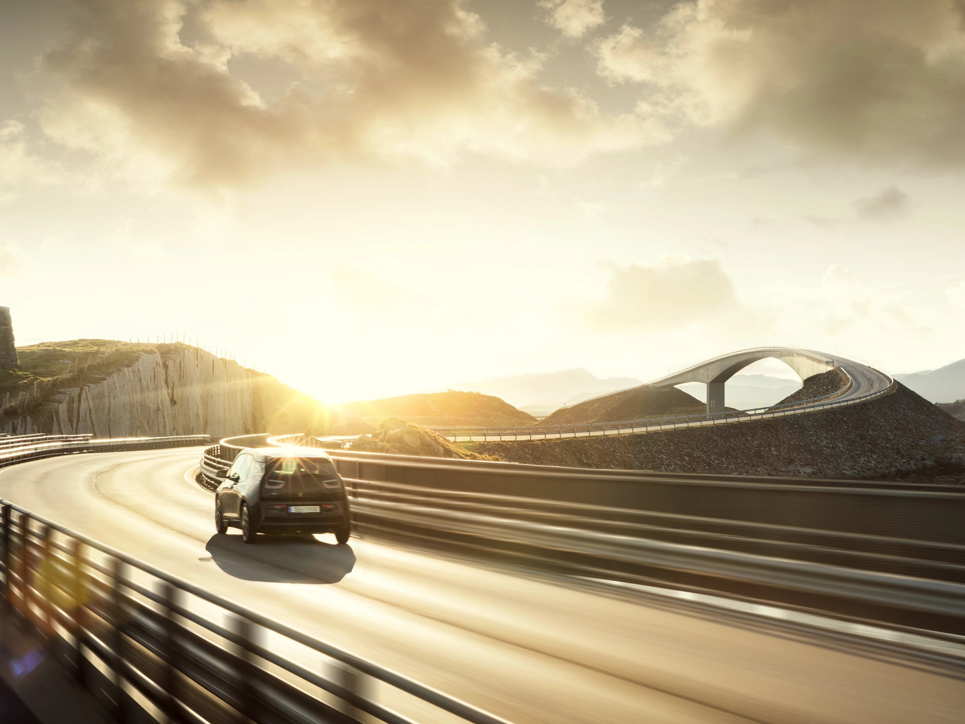 A car drives across a curving bridge at sunset in a coastal landscape.