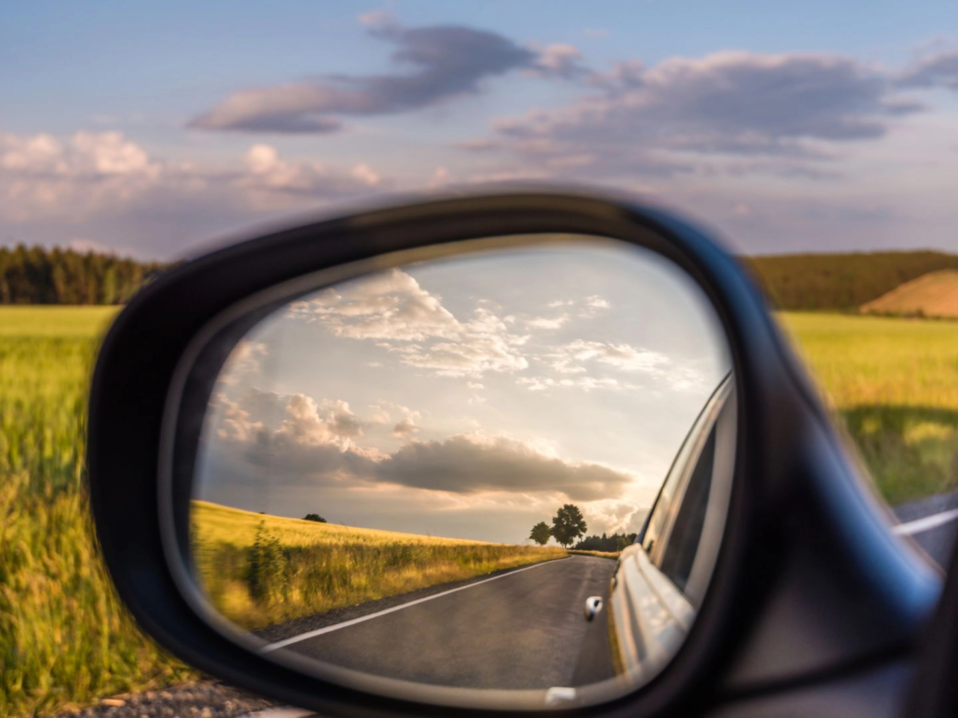 Landstraße und bewölkter Himmel spiegeln sich im Seitenspiegel eines Autos bei Tageslicht.