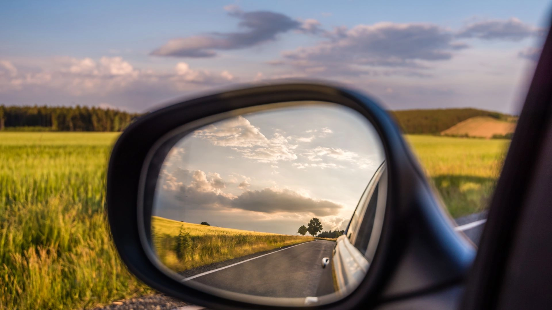 Landstraße und bewölkter Himmel spiegeln sich im Seitenspiegel eines Autos bei Tageslicht.