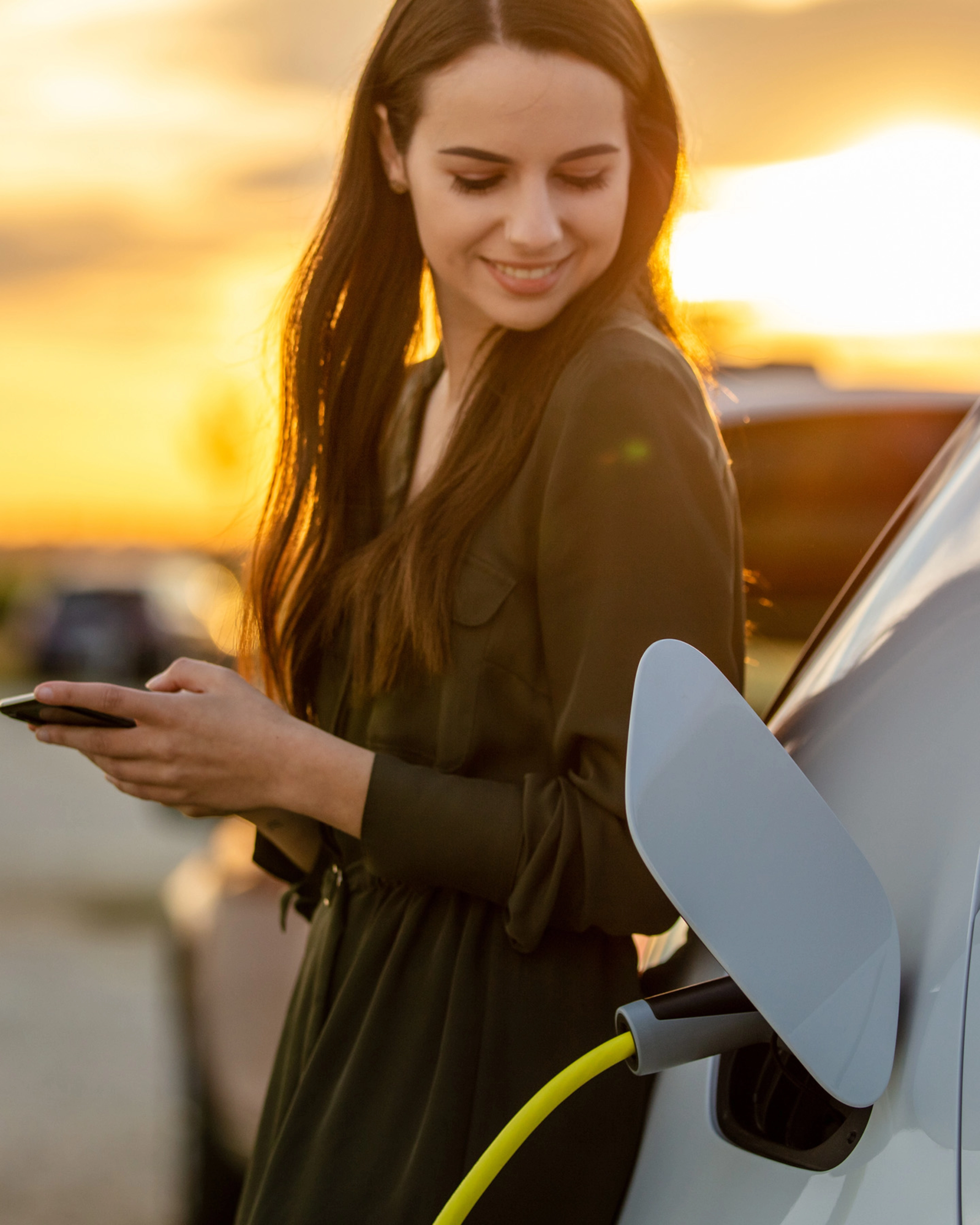 Eine Frau lächelt bei Sonnenuntergang, während ihr E-Auto lädt und sie ein Smartphone hält.