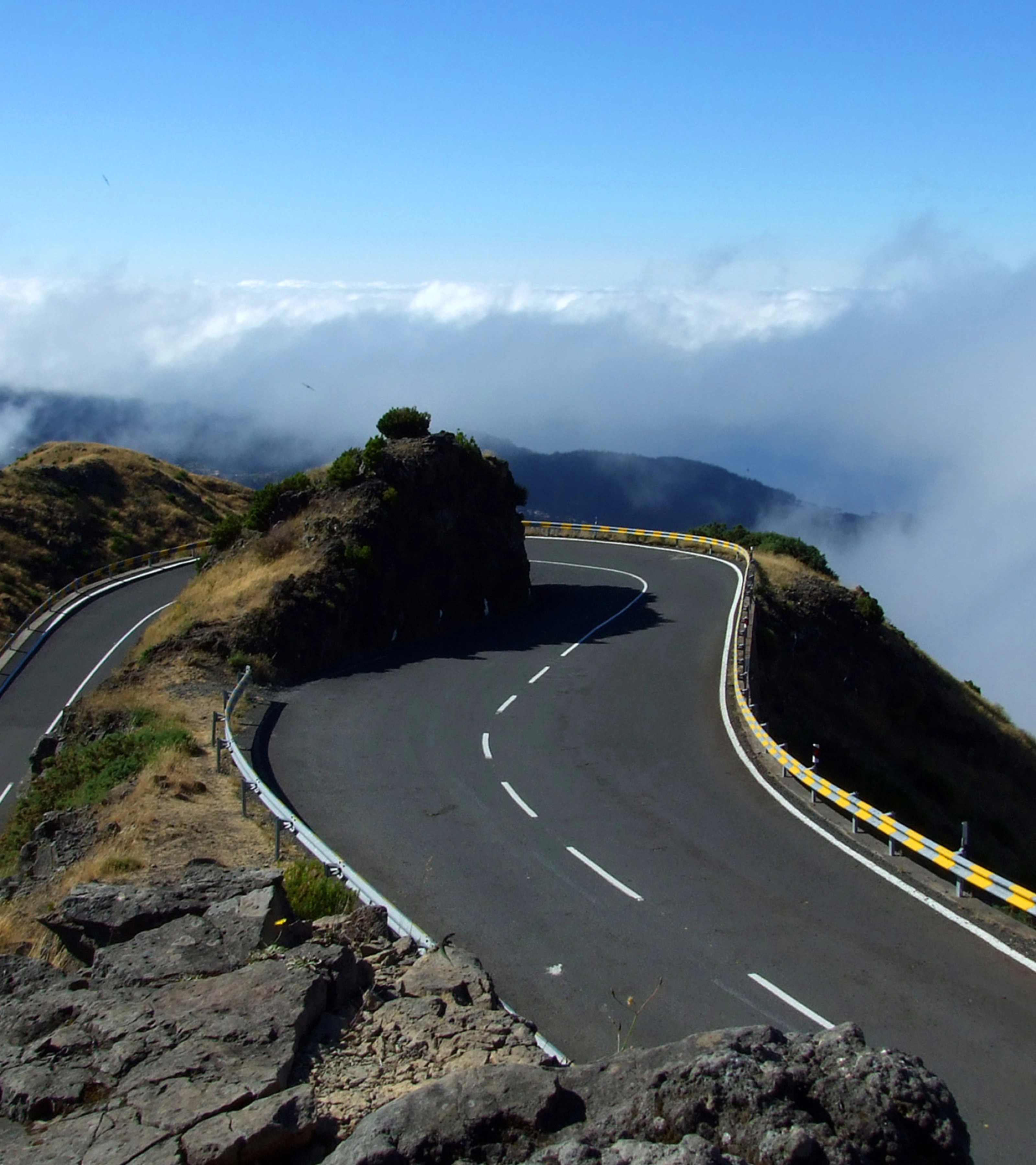 Kurvige Bergstraße mit Leitplanken und nebligem Wolkenhintergrund.