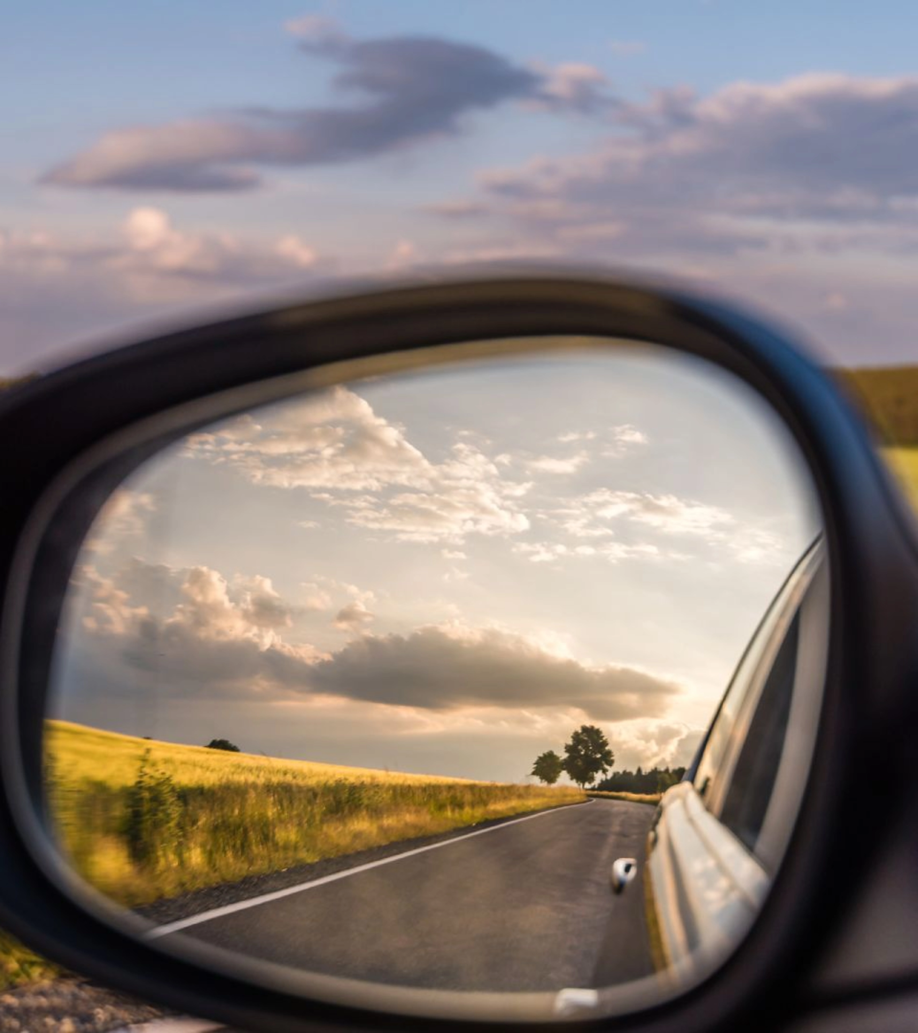Landstraße und bewölkter Himmel spiegeln sich im Seitenspiegel eines Autos bei Tageslicht.