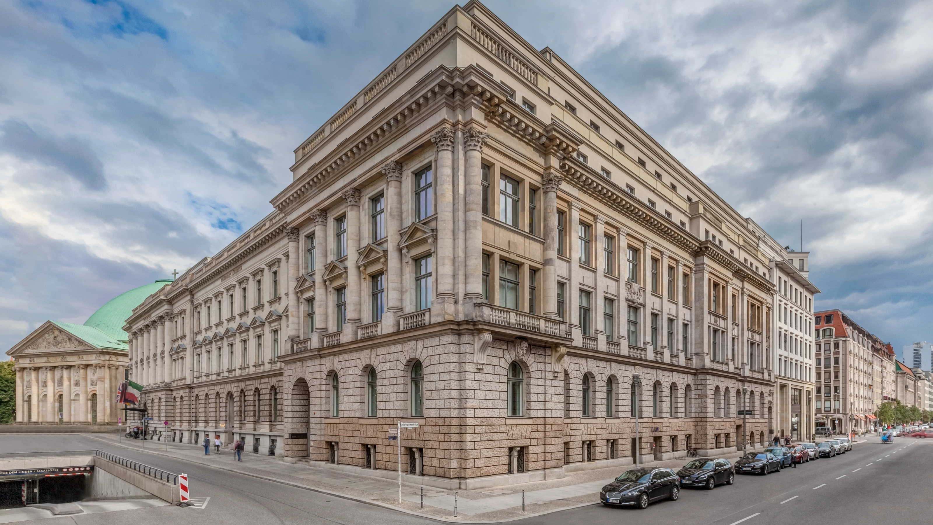 Corner view of a large neoclassical building with columns and parked cars along the street.