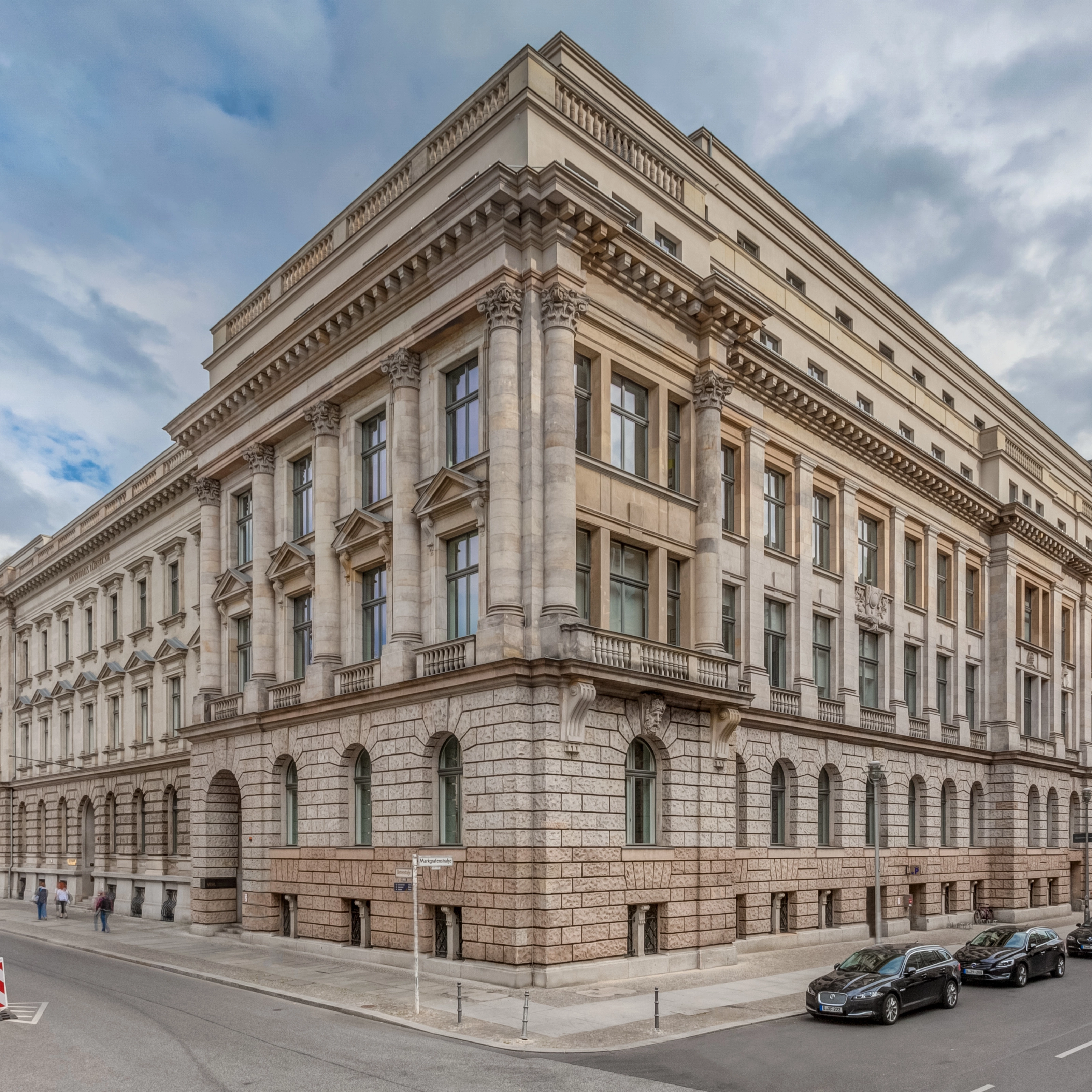 Corner view of a large neoclassical building with columns and parked cars along the street.
