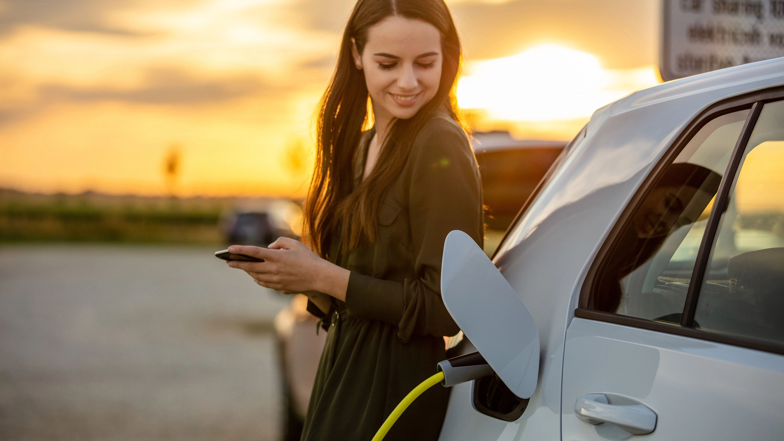 Eine Frau lächelt bei Sonnenuntergang, während ihr E-Auto lädt und sie ein Smartphone hält.