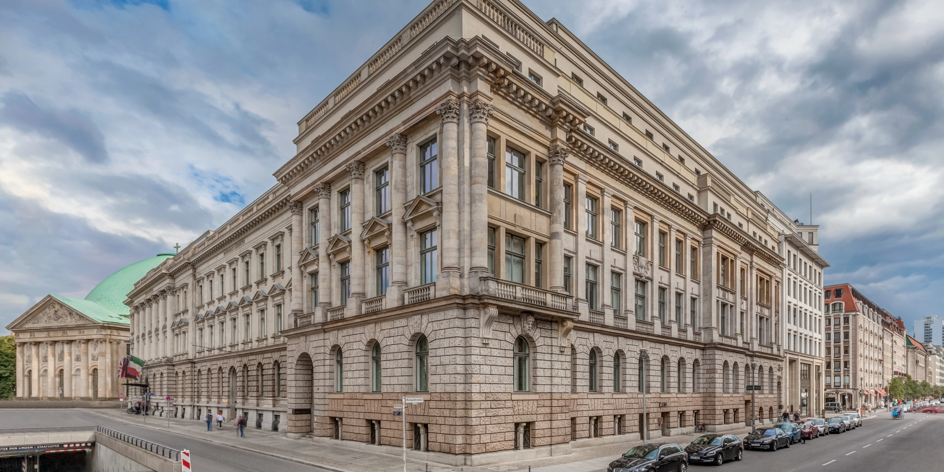 Corner view of a large neoclassical building with columns and parked cars along the street.