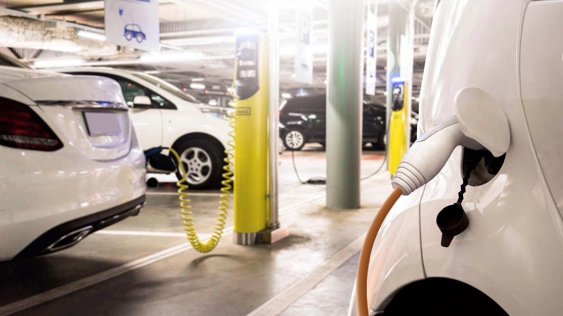 Electric cars charging at a yellow charging station in an indoor parking garage.