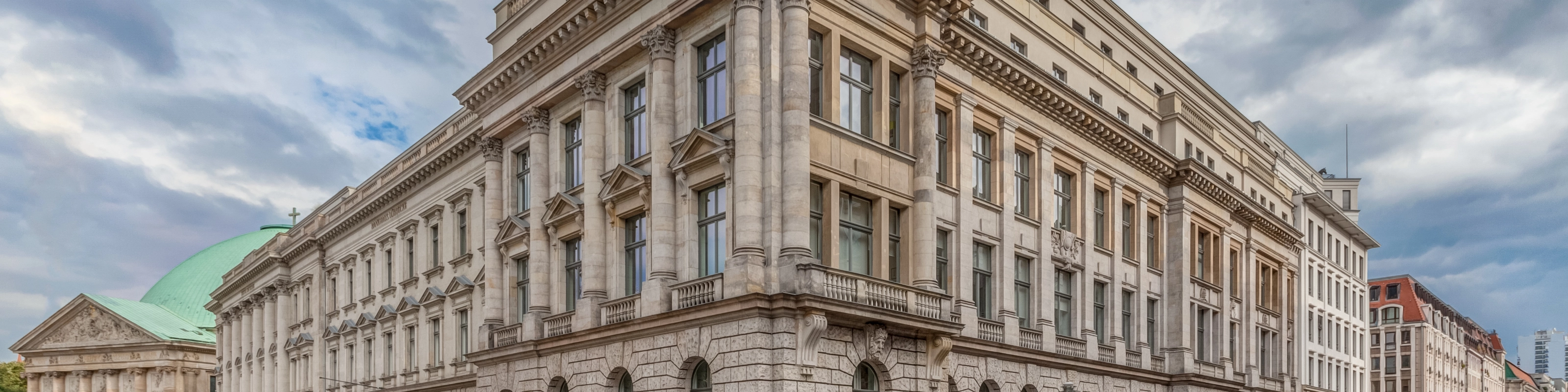 Corner view of a large neoclassical building with columns and parked cars along the street.
