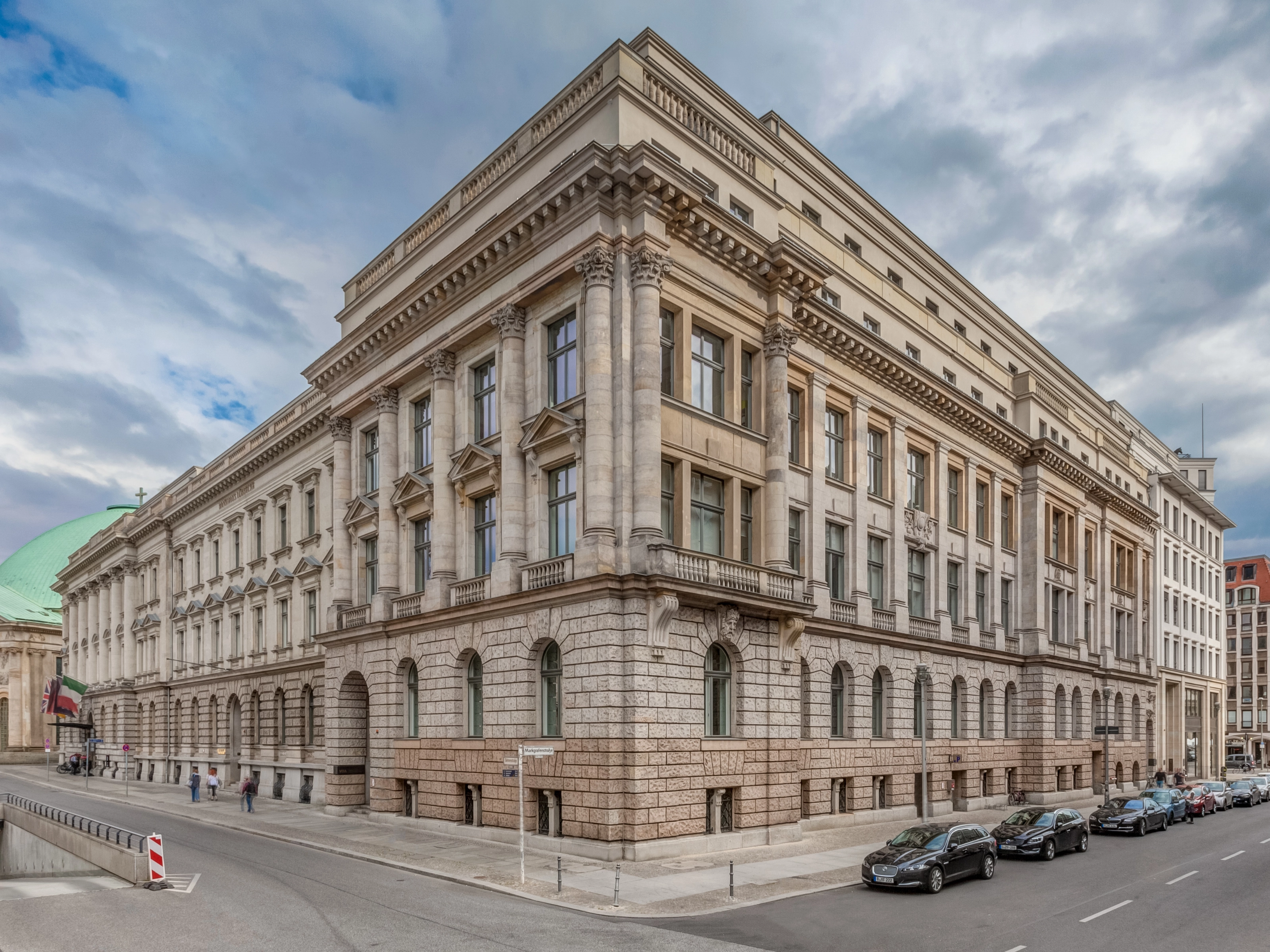 Corner view of a large neoclassical building with columns and parked cars along the street.