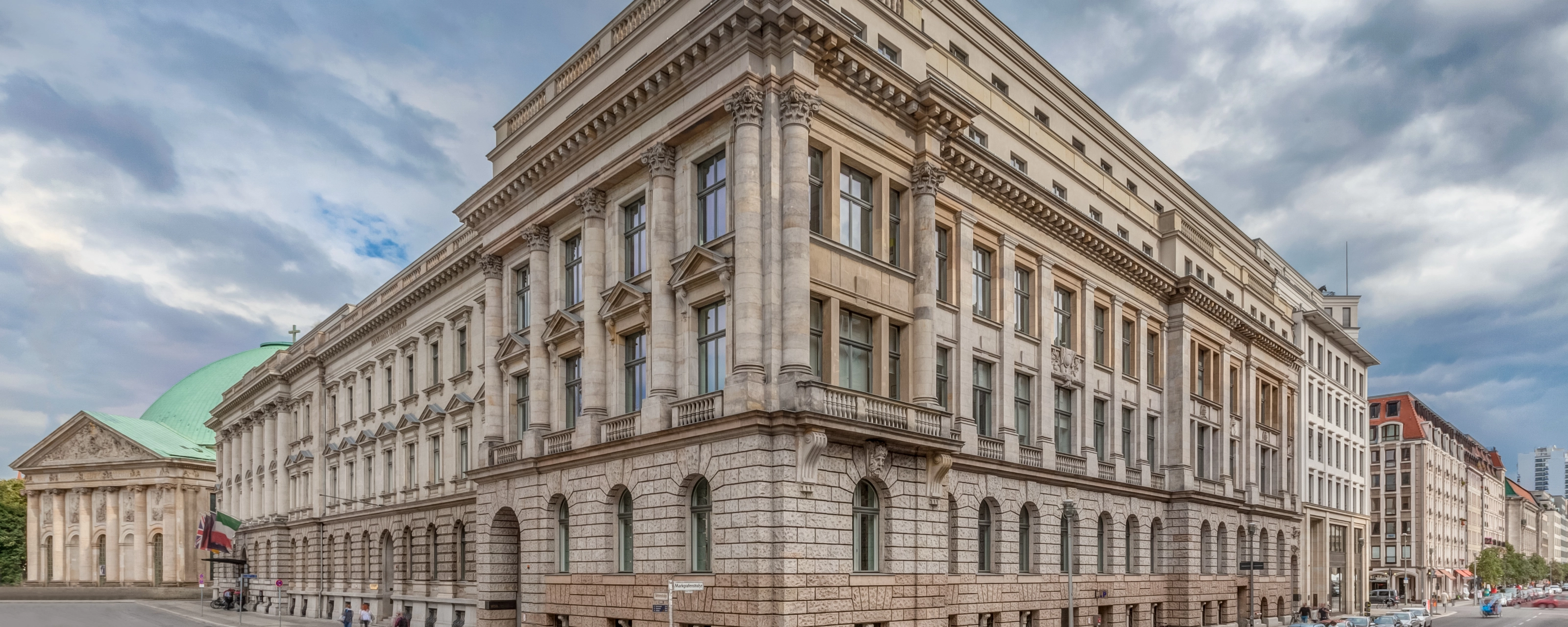 Corner view of a large neoclassical building with columns and parked cars along the street.