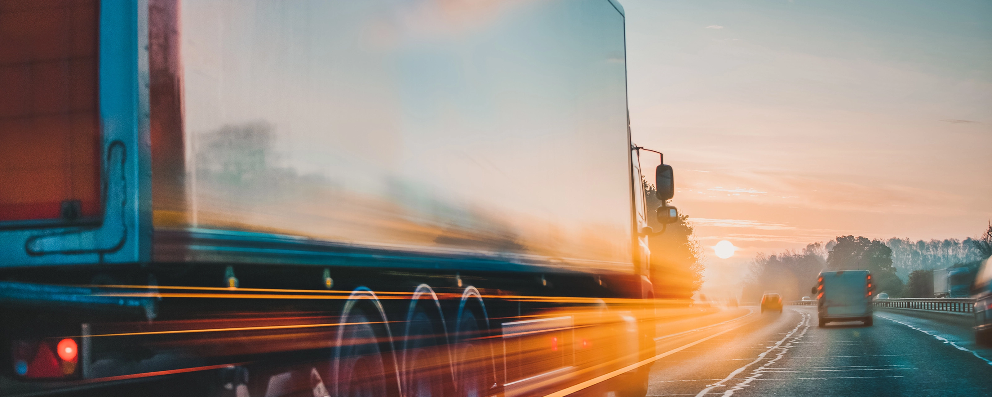 Lkw fährt bei Sonnenaufgang mit Lichtspuren und Bewegungsunschärfe auf der Autobahn.