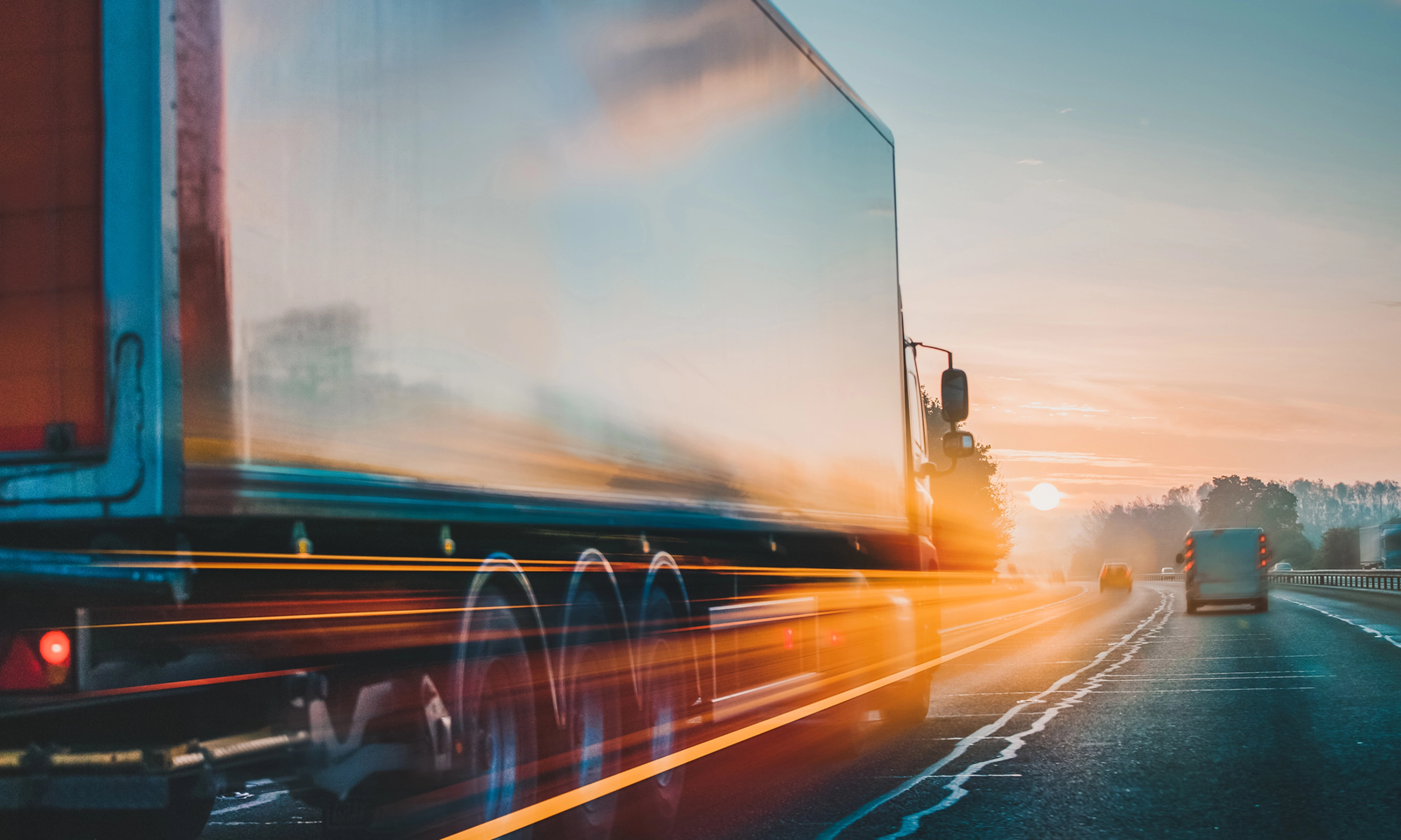 Lkw fährt bei Sonnenaufgang mit Lichtspuren und Bewegungsunschärfe auf der Autobahn.