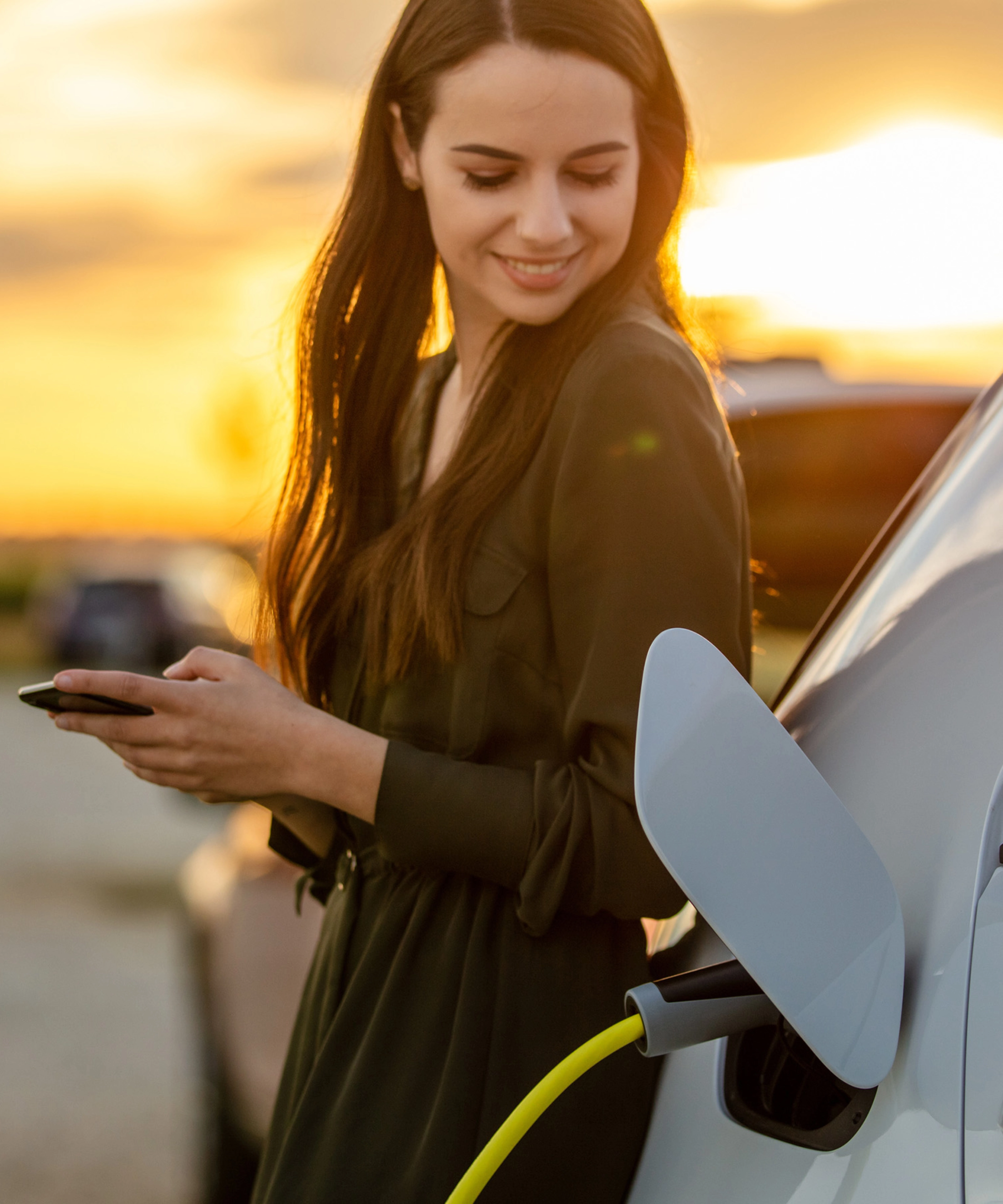 Eine Frau lächelt bei Sonnenuntergang, während ihr E-Auto lädt und sie ein Smartphone hält.