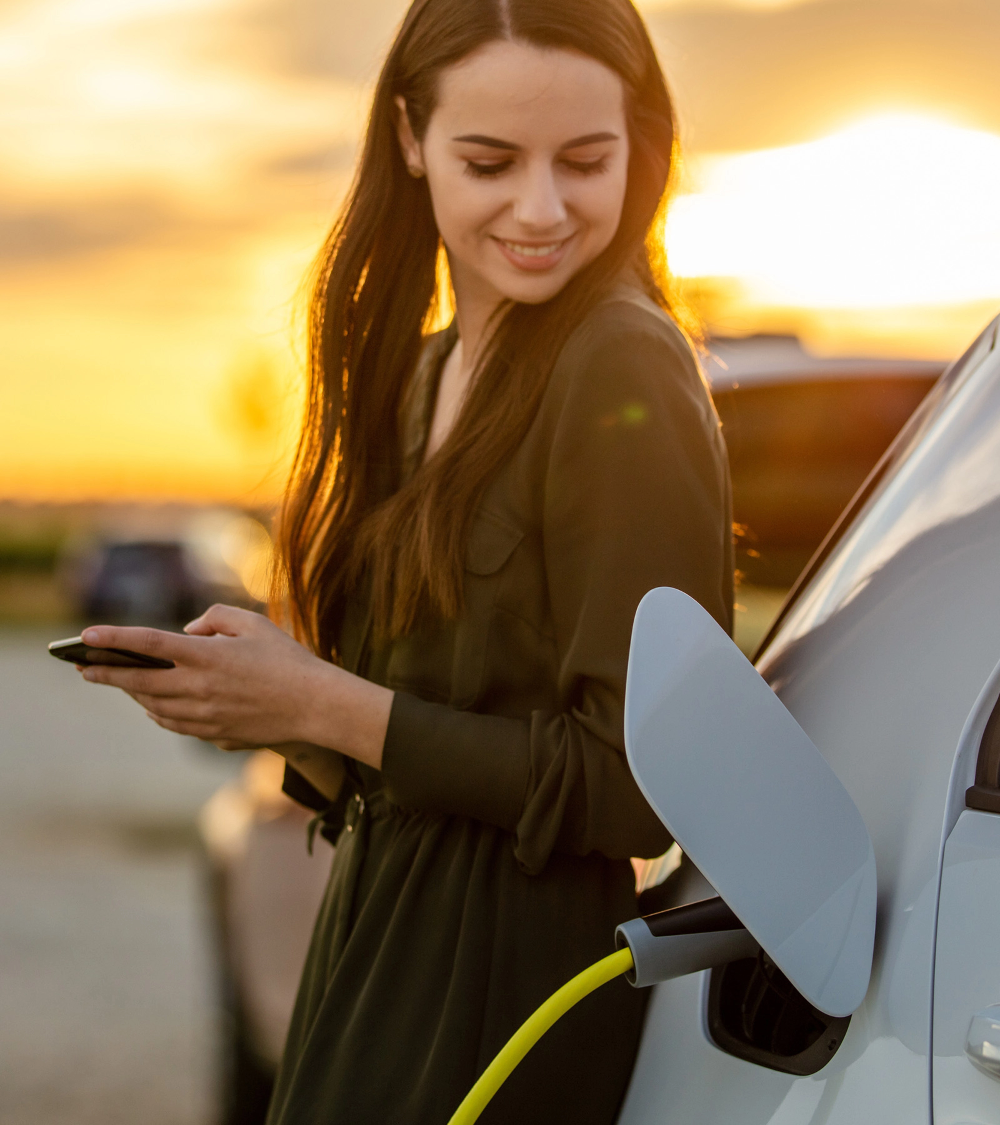 Eine Frau lächelt bei Sonnenuntergang, während ihr E-Auto lädt und sie ein Smartphone hält.